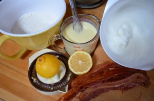 bowl of dry ingredients, measuring glass of wet ingredients, whipped egg white in a separate bowl, lemon and lemon juicer and strips of cooked bacon