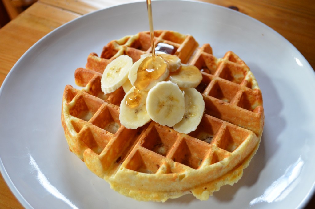a white plate with one waffle on it. sliced bananas on top and maple syrup being poured on