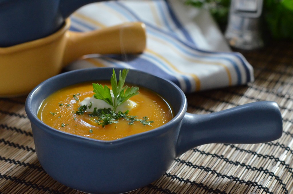 butternut squash soup in a blue crock with sour cream and parsley. Yellow crock and tea towel in background.