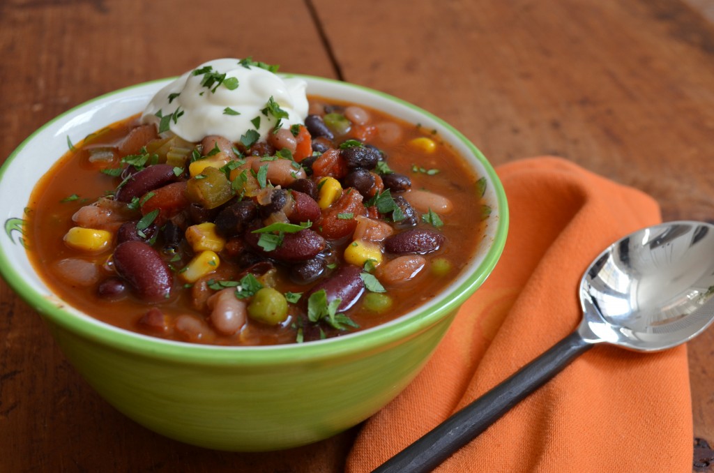 chili in a green bowl on a wooden table with an orange napkin beside it.