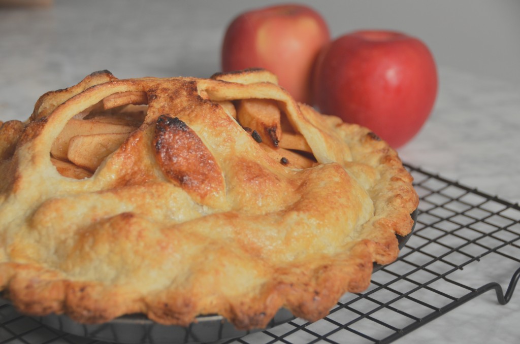An apple pie on a cooling rack with two red apples in the background