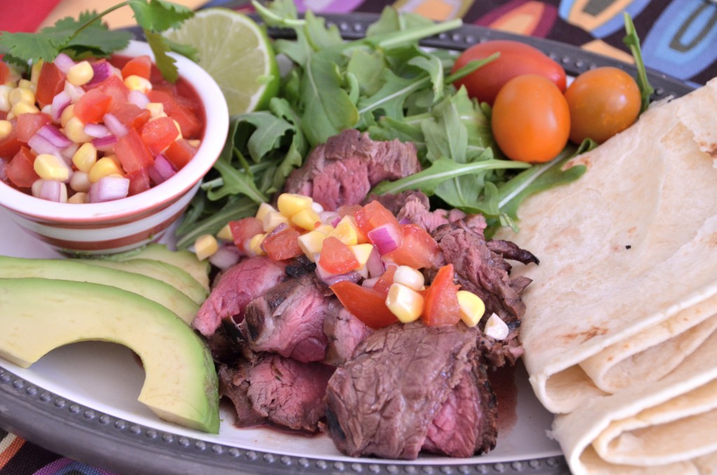 A white plate with corn and tomato salsa in a small bowl, sliced avocado, sliced beef with some salsa on top and folded flour tortillas