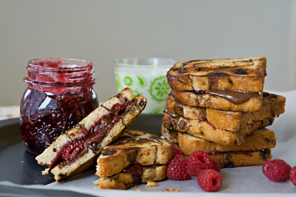 a stack of sandwiches on a plate, one cut open in half, a pot of jam and a glass of milk