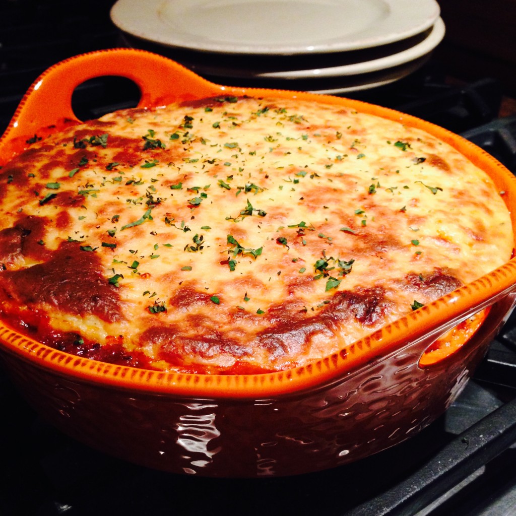 A large round orange ceramic dish with Moussaka in it. A stack of plates in the background